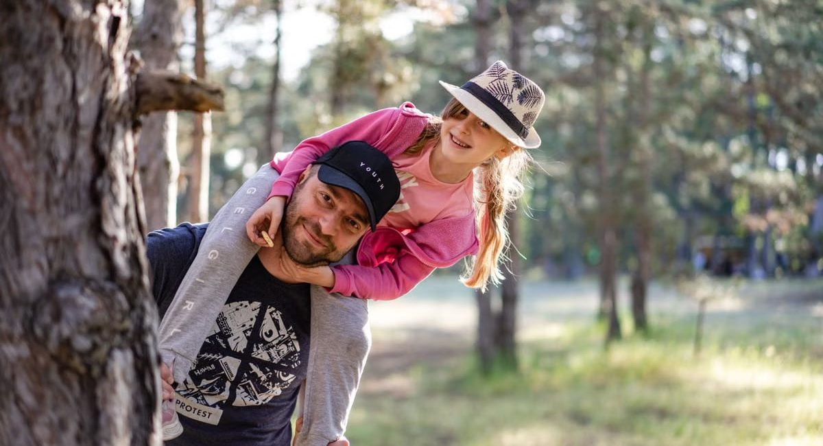 A joyful father giving his daughter a piggyback ride in a sunny forest setting.