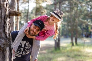 A joyful father giving his daughter a piggyback ride in a sunny forest setting.